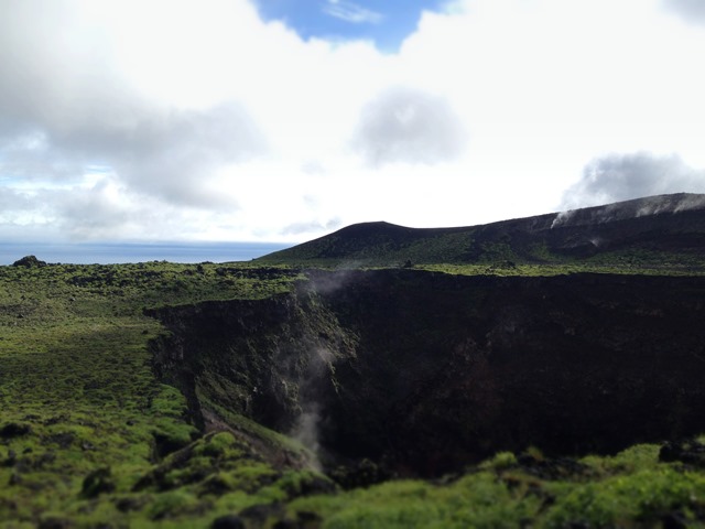 Green grassy volcanic crater landscape with steam rising under a cloudy sky, with a distant ocean horizon in calm natural tones